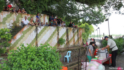People sitting below the huge trees along the river to shade away from the heat.